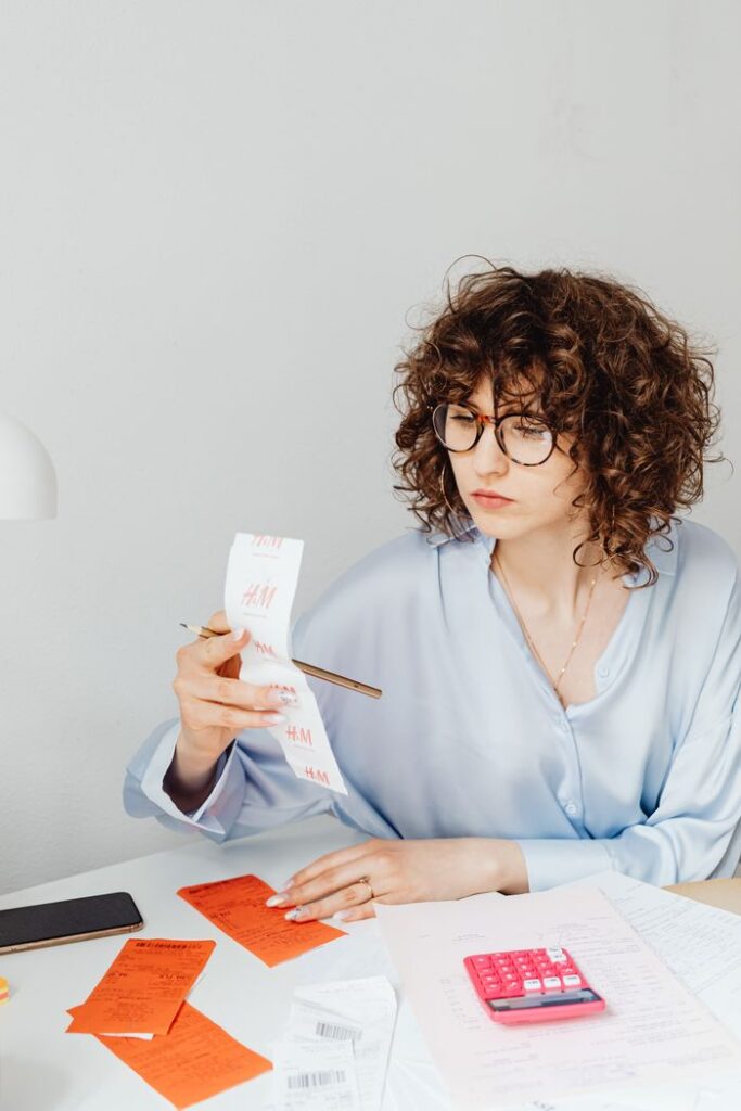 Curly-haired woman examining receipts with a calculator at her desk.